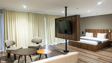 Habitación de hotel con cama, televisión montada en un panel de madera, zona de estar con una silla y una mesita, y una ventana grande con cortinas.
