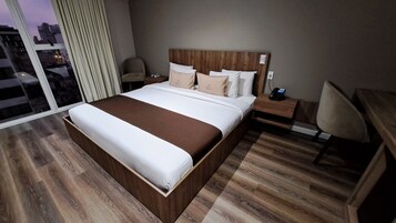 Habitación de hotel con una cama grande, cabecera de madera, mesita de noche con teléfono y una silla.