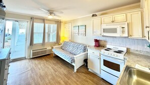 Une cuisine compacte équipée d’une cuisinière intégrée, d’un four à micro-ondes et d’un évier. On y trouve une fenêtre avec des stores, un ventilateur de plafond et un petit coin salon avec un coussin à motifs.