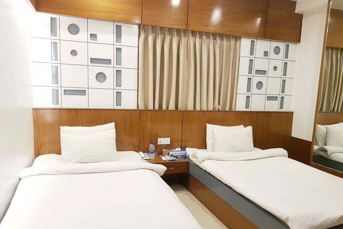 A hotel room with two beds, wooden headboards, a mirror, a bedside table with a phone and a bottle, and a curtain with a geometric pattern.