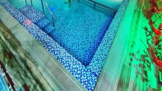 Ein überdachter Schwimmbereich mit gefliestem Boden, umgeben von einem Geländer, beleuchtet durch farbige Lichter.