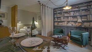 Une pièce chaleureuse avec une table à manger dressée pour deux personnes, une bibliothèque remplie de livres, un fauteuil vert et une valise ancienne.