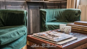 Une pièce avec deux fauteuils verts, une table en bois sur laquelle sont posés des livres et une théière, et une cheminée en arrière-plan.