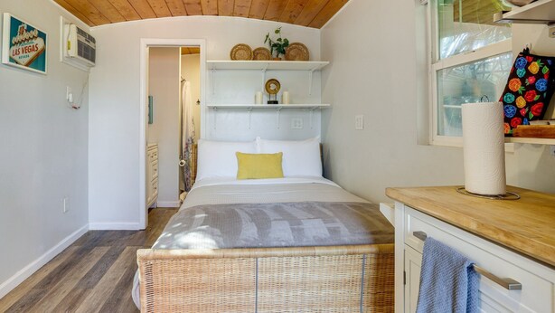 A bedroom with a bed, a wicker bench, a window with a towel, and a shelf with decorative items. - Private Tiny House Retreat with Spacious Patio, Free Parking & WiFi (Las Vegas)