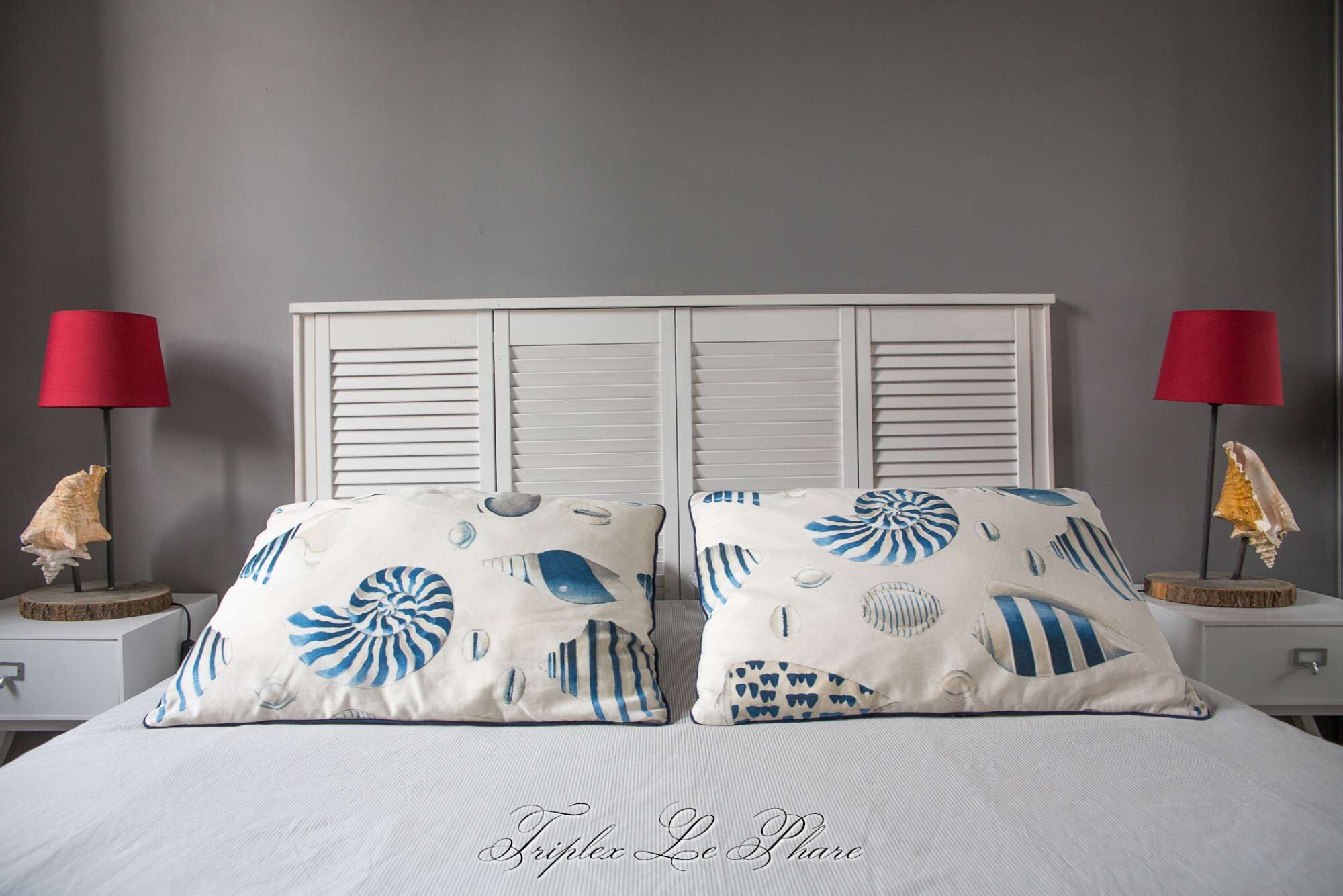Un lit avec deux oreillers ornés de motifs marins, flanqué de deux tables de chevet avec des lampes rouges et des coquillages.