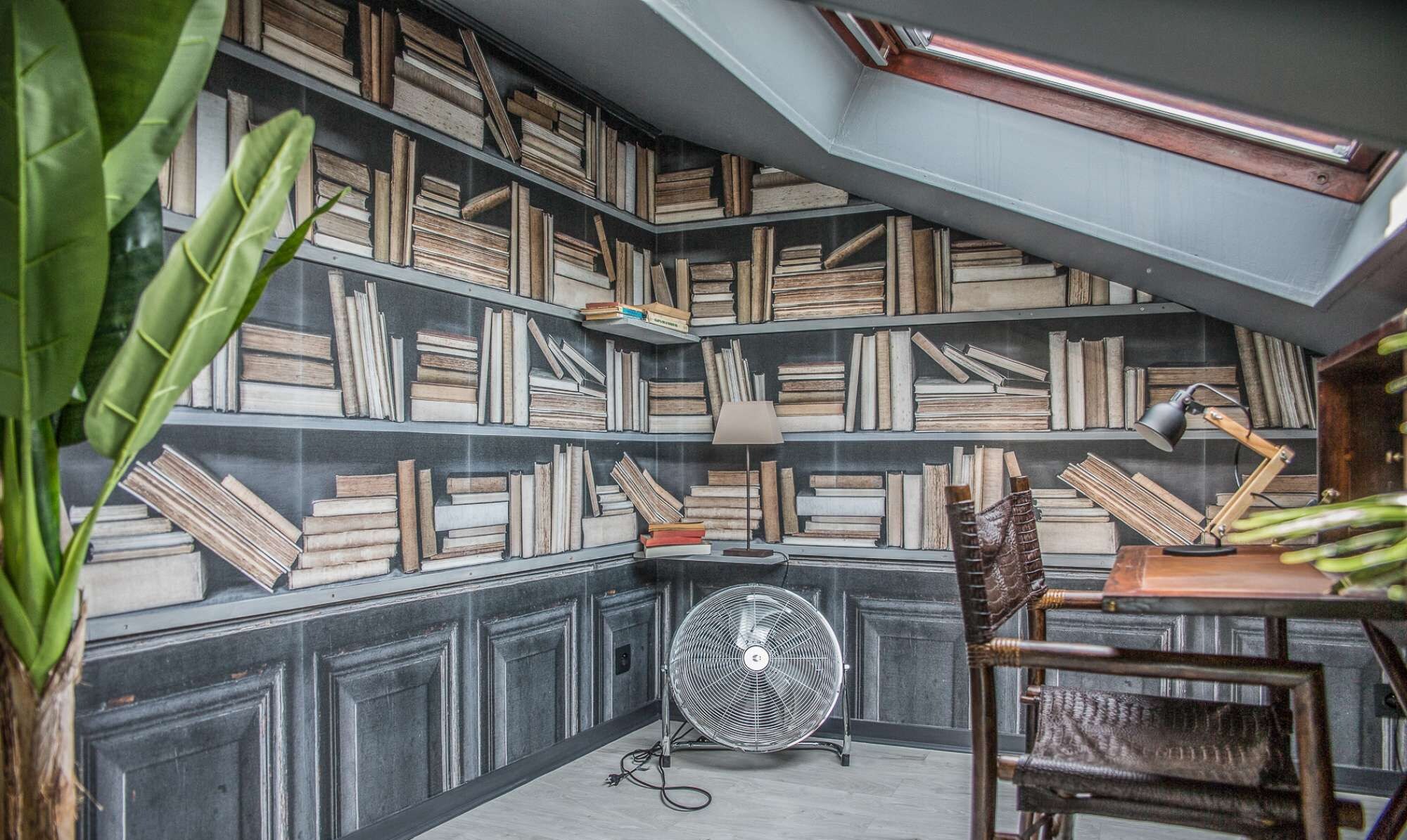 A cozy reading nook with a bookshelf filled with books, a fan, and a chair.