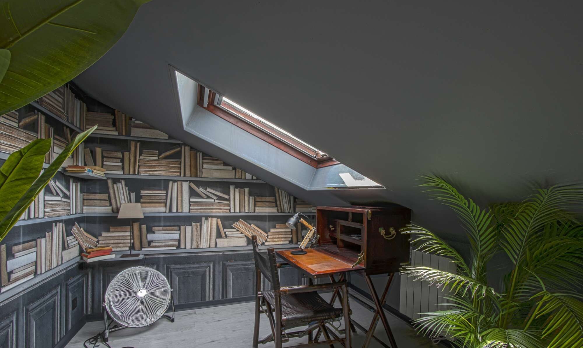 Ein Zimmer mit schrägem Dach, einem Oberlicht, einem Holztisch, einem Stuhl, einem Ventilator und einem Bücherregal voller Bücher.