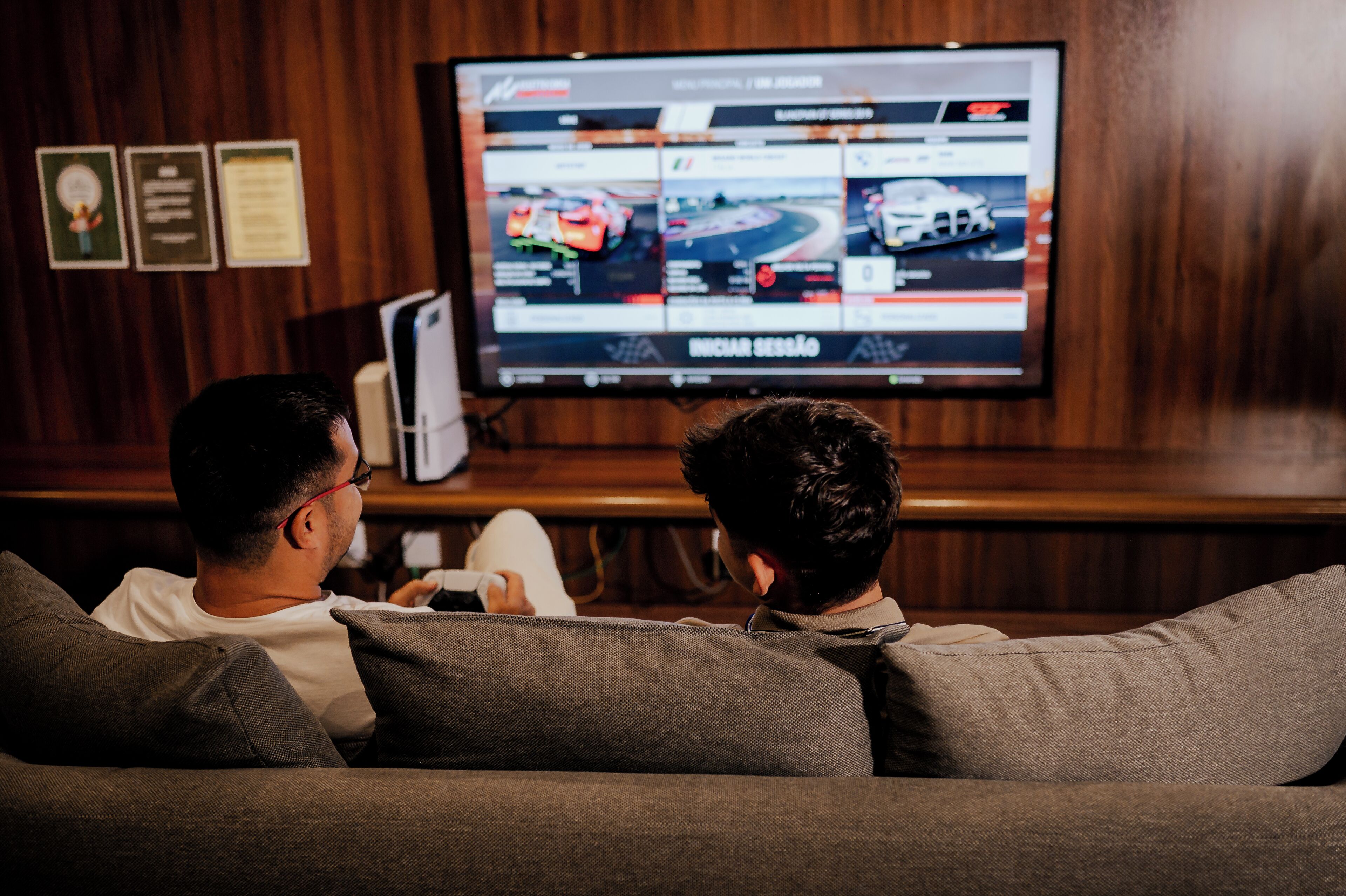 Two people watching a racing video on a large TV screen in a room with wooden paneling.