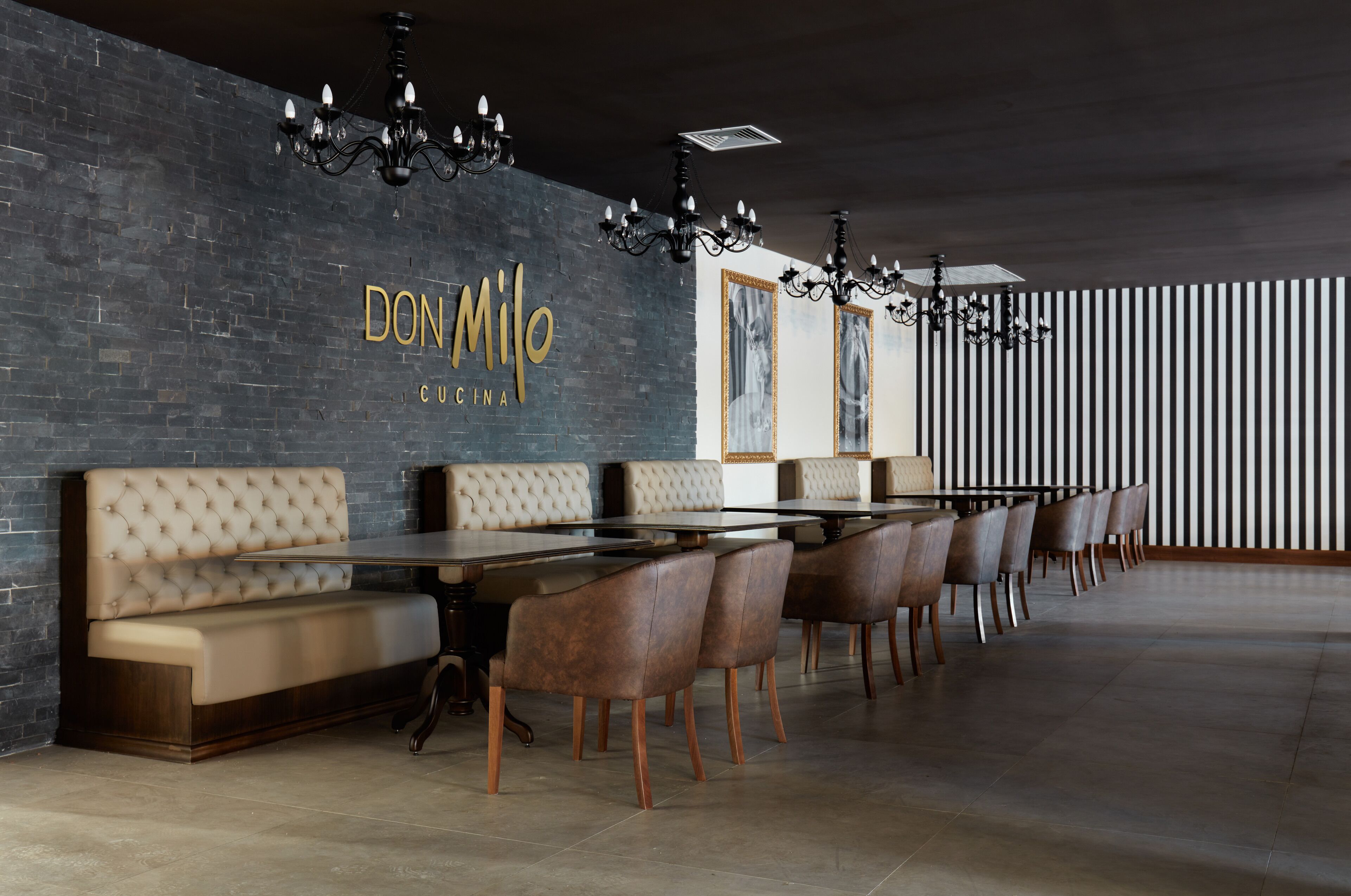 A modern restaurant interior with a long dining table, cushioned benches, and a dark brick wall with the name 'DON MILO CU CINA'.