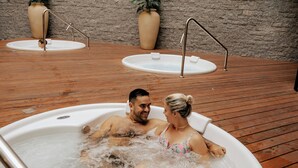 Una pareja en un jacuzzi con una terraza de madera y dos sillones de spa al fondo.