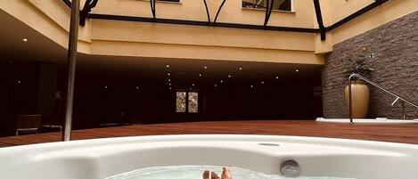 A hot tub in a multi-level indoor pool area with a modern architectural design, including a unique ceiling structure and a stone wall.