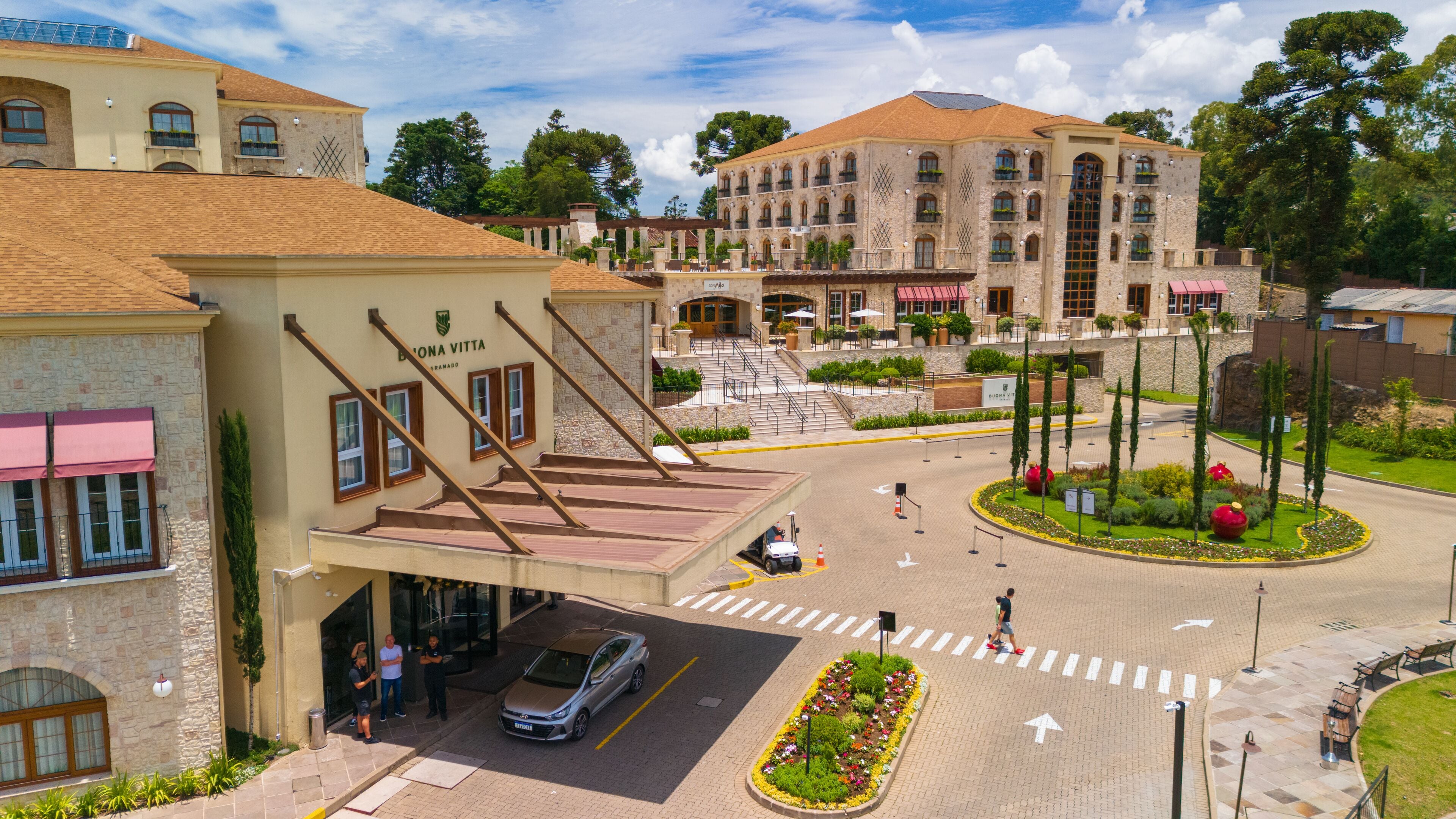 Un hotel con una escalera destacada que conduce a la entrada, un área de estacionamiento y un jardín ajardinado con un parterre circular.