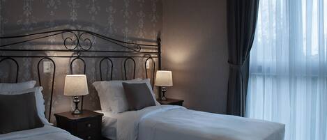 Habitación de hotel con dos camas, cada una con mesita de noche, lámparas y almohadas. La habitación tiene papel tapiz estampado y un ventanal grande con cortinas.