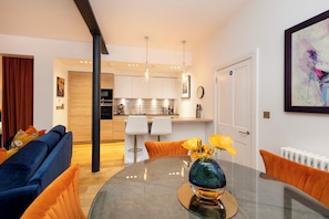 A modern living room with a glass dining table, a sofa, and a kitchen in the background.