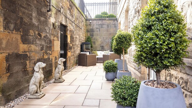 A courtyard with stone walls, a seating area with cushions, and two potted trees. - Edinburgh Luxury Apartment- The Matisse Suite (Edinburgh)