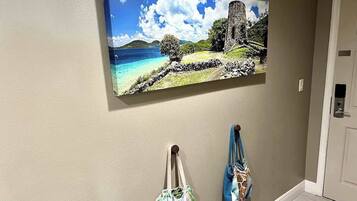 A hallway with a painting of a coastal landscape, a door, and two bags hanging on hooks.