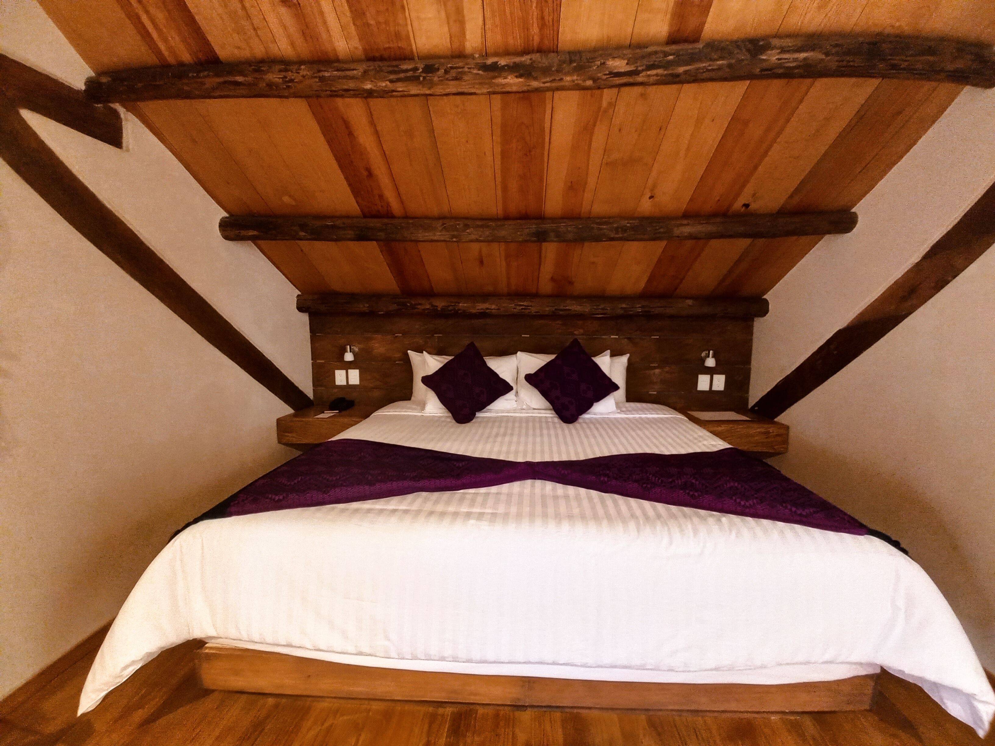 A bedroom with a wooden ceiling, a bed with white bedding and purple pillows, and a wooden headboard.