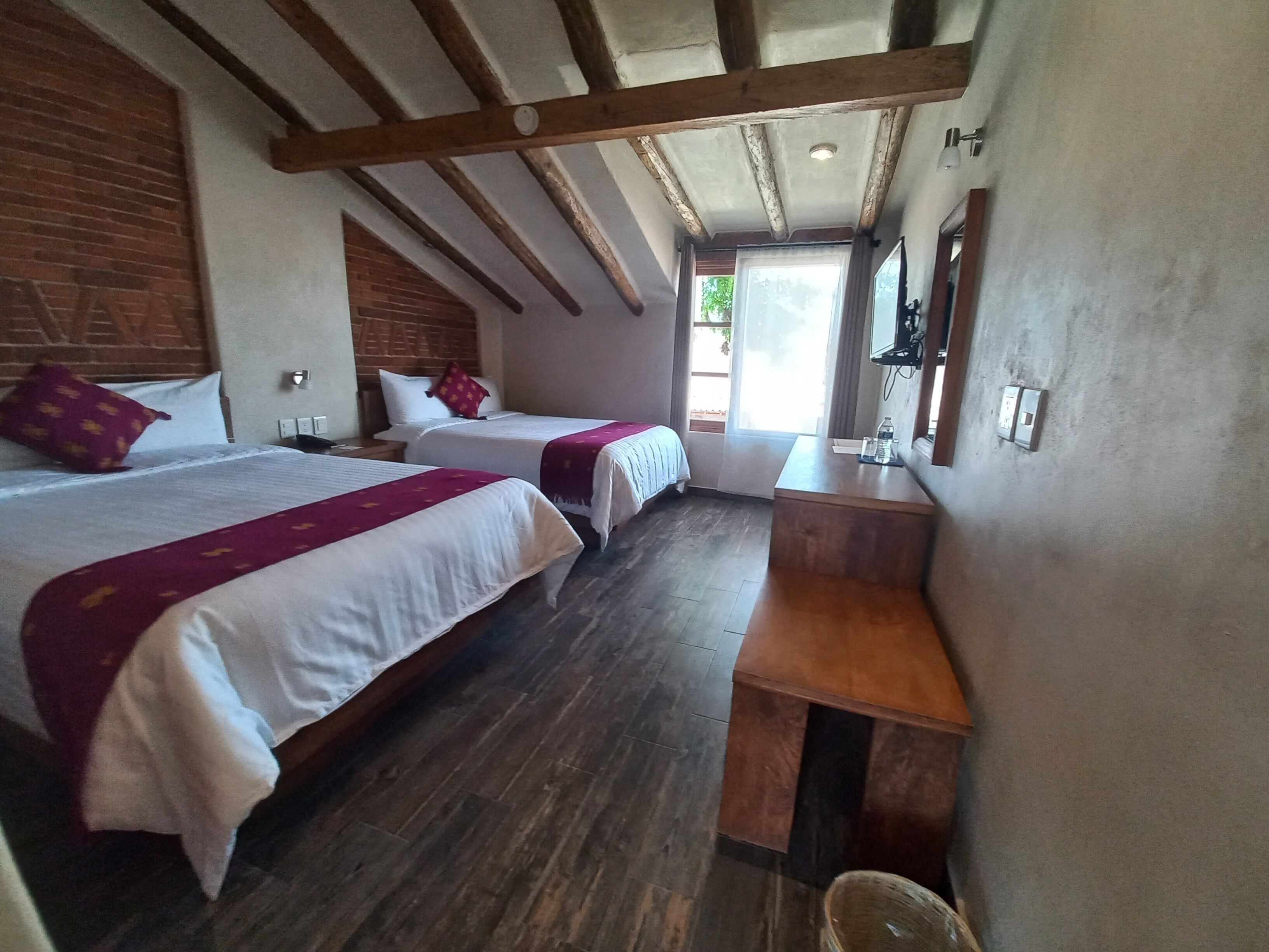 A hotel room with two beds, wooden beams on the ceiling, a small wooden desk, and a window with a view.