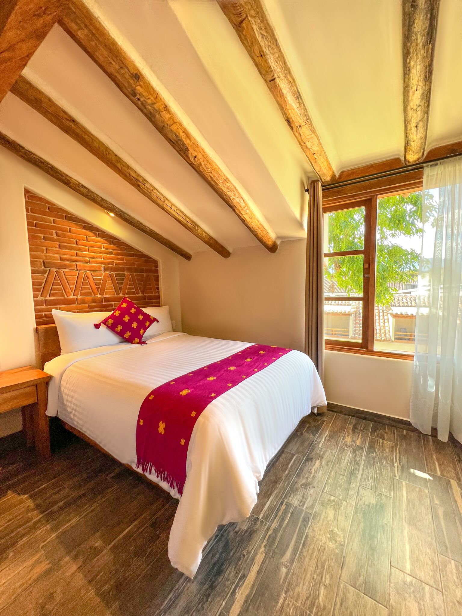 A bedroom with a wooden ceiling, a brick wall, a large window with curtains, and a bed with a white bedspread and a pink blanket.