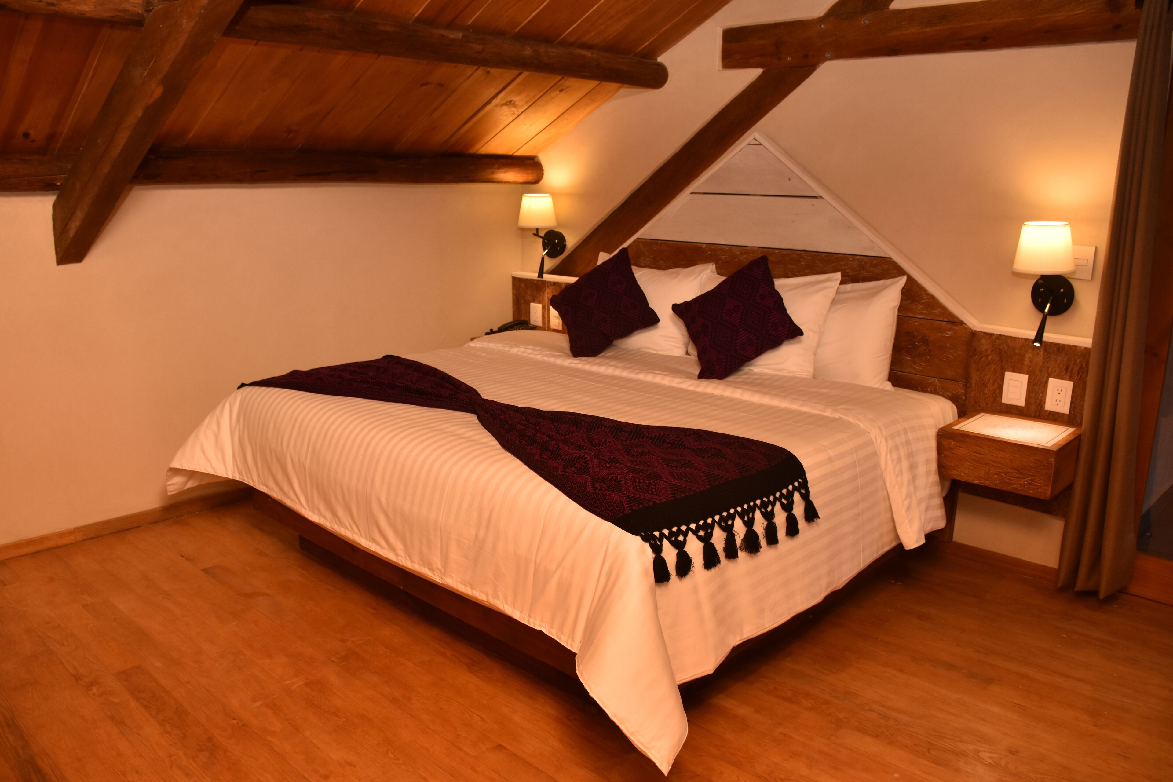 A bedroom with a wooden ceiling, a large bed with white bedding and dark pillows, two bedside lamps, and a wooden nightstand.