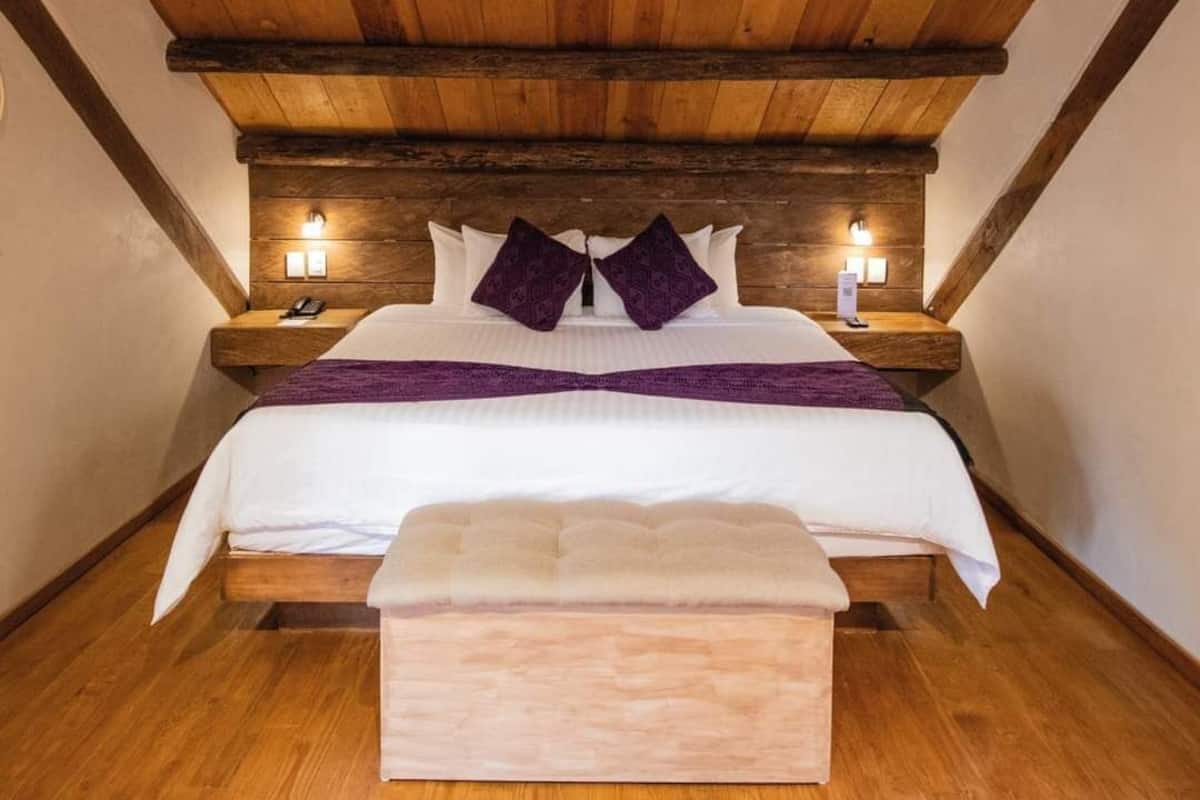 Un dormitorio con techo de madera, cama con ropa de cama blanca y almohadas moradas, lámparas de noche y un banco de madera al pie de la cama.