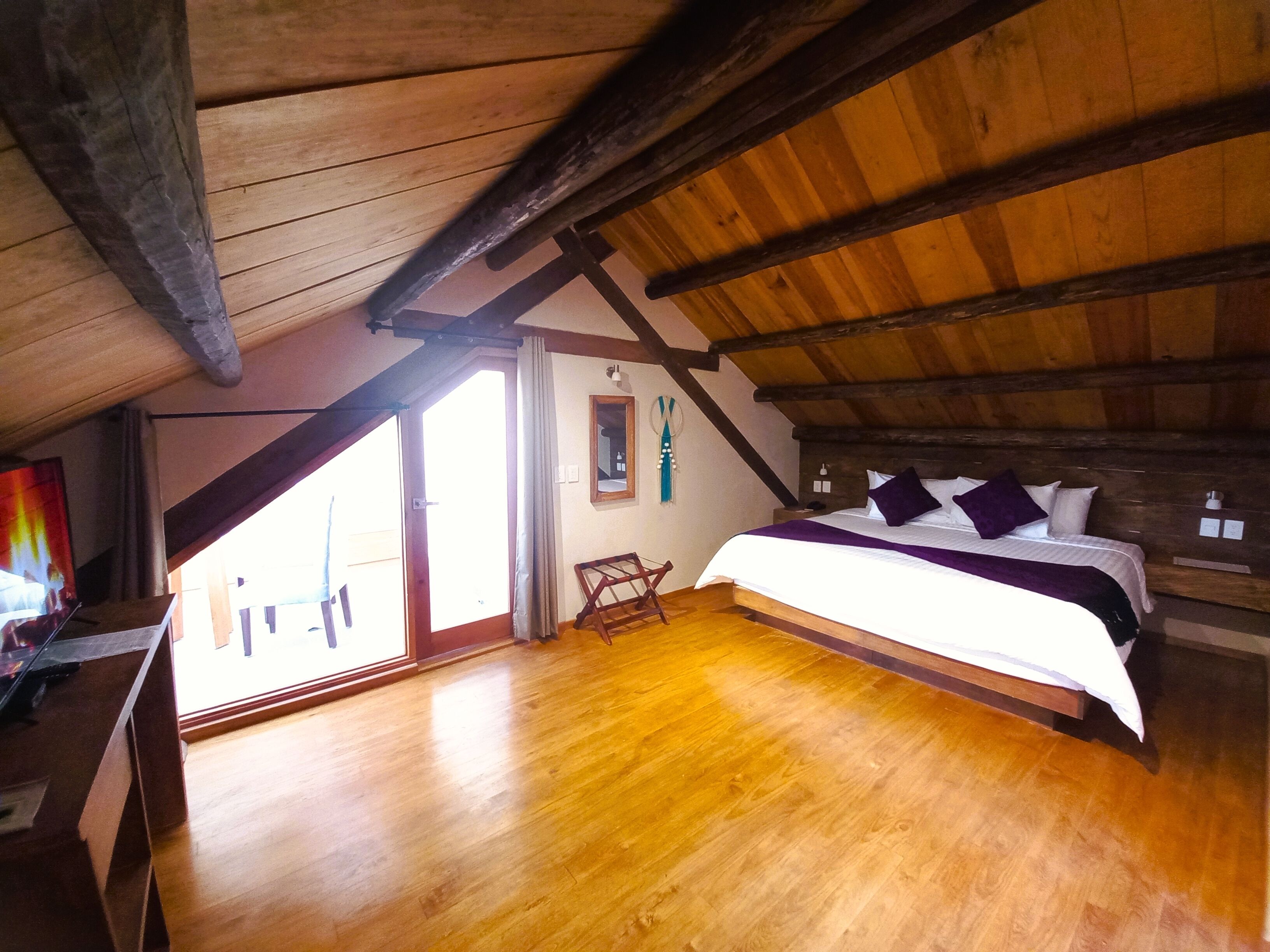 Une chambre avec un plafond en bois, un lit recouvert d’une literie blanche et violette, et une vue dégagée sur l’extérieur grâce à une grande fenêtre.