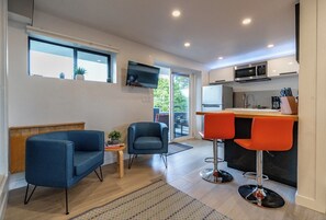 En kompakt opholdsstue med tekøkken, spiseplads med to stole, sofa og tv ophængt på væggen.