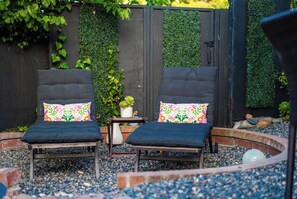 To mørke loungesenge med farverige blomstermønstrede puder, placeret på en grusoverflade med et lille bord og en potteplante imellem.