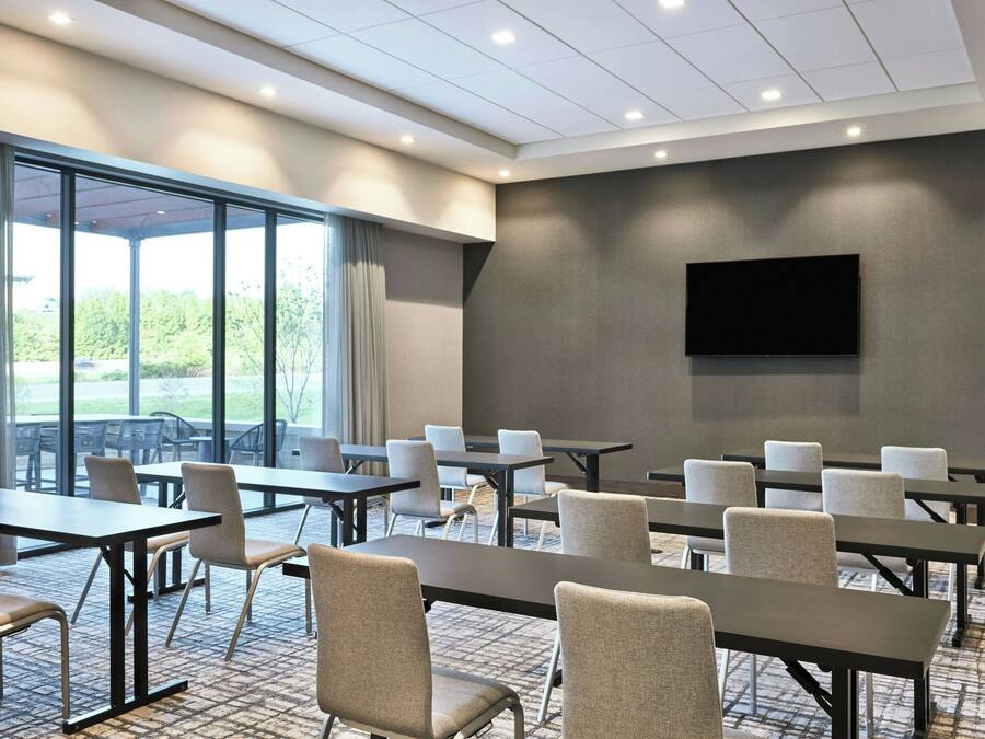Une salle de conférence équipée de tables et de chaises, de grandes fenêtres et d’un téléviseur à écran plat fixé au mur.