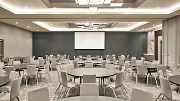 Sala de conferência com várias mesas e cadeiras, uma tela grande e luminárias modernas.