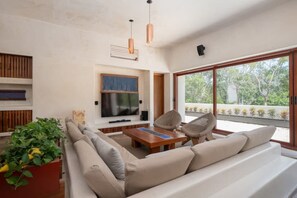 Ein modernes Wohnzimmer mit einem großen Sofa, einem Couchtisch und zwei Sesseln. An der Wand ist ein Flachbildfernseher befestigt, es gibt einen Blumentopf und ein Fenster mit Blick auf grüne Natur.