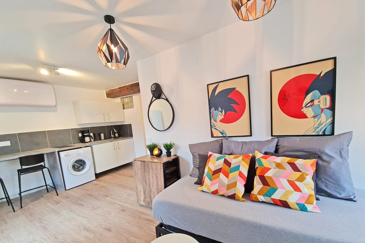 A modern apartment with a kitchenette, a washing machine, a bed with colorful pillows, and two abstract paintings.
