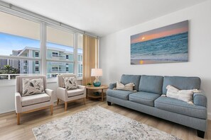 Ein Wohnzimmer mit einer Couch, zwei Sesseln, einem Couchtisch und einem Gemälde, das einen Sonnenuntergang über dem Meer zeigt.