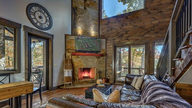 A cozy living room with a fireplace, leather sofa, and a large clock on the wall. - Singing Pines - Mountain Views | Hot Tub | Screened Porch | Fire Pit (Blue Ridge)