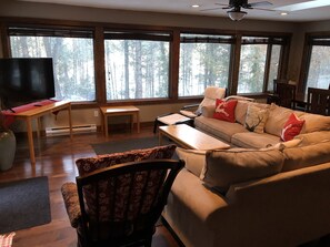 A living room with a sofa, a coffee table, a TV, and large windows offering a view of trees.