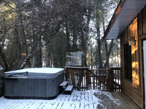 A wooden cabin with a hot tub on a deck surrounded by trees.