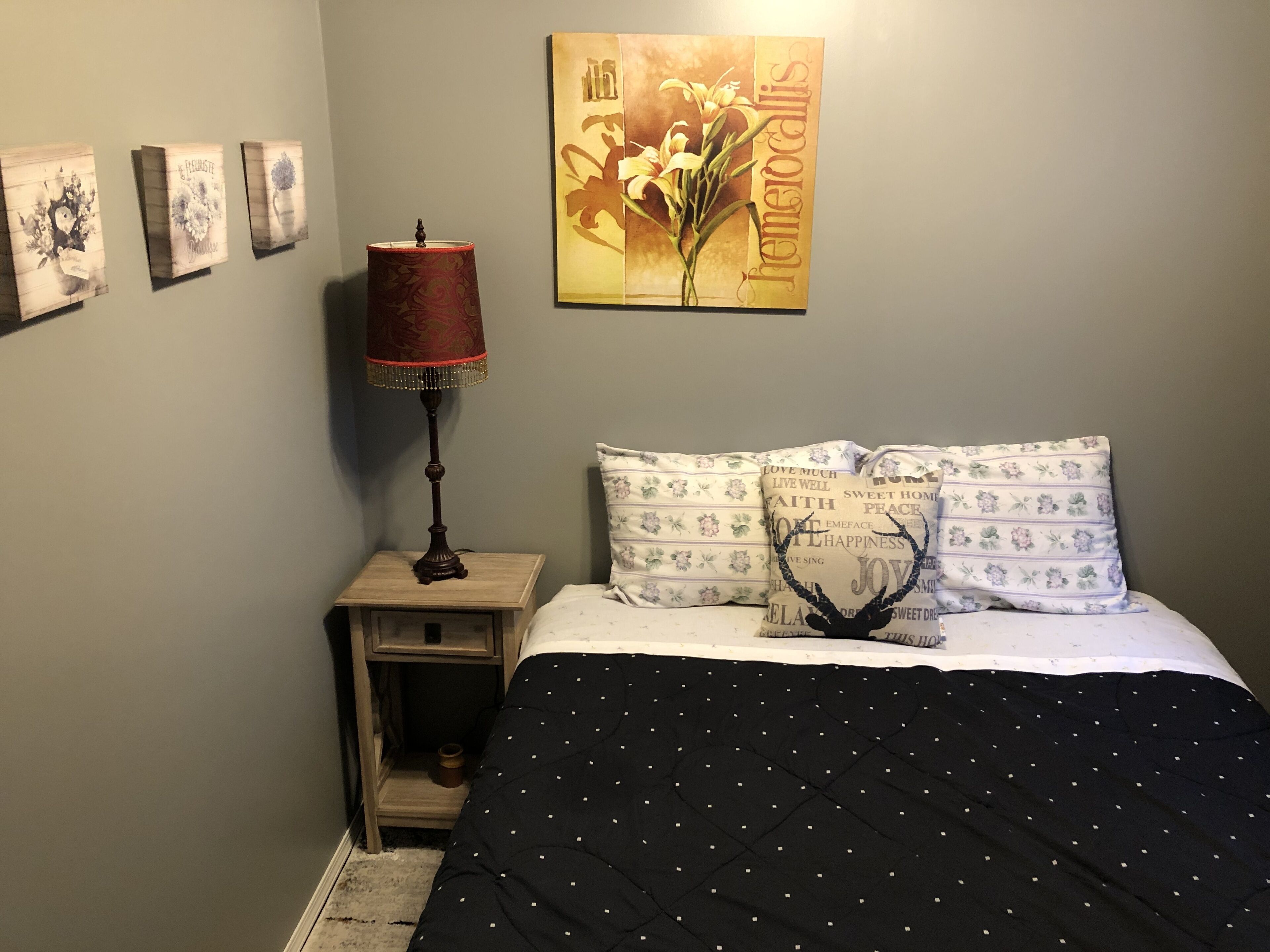 Un dormitorio con cama, mesita de noche, lámpara y una pared decorada con marcos.
