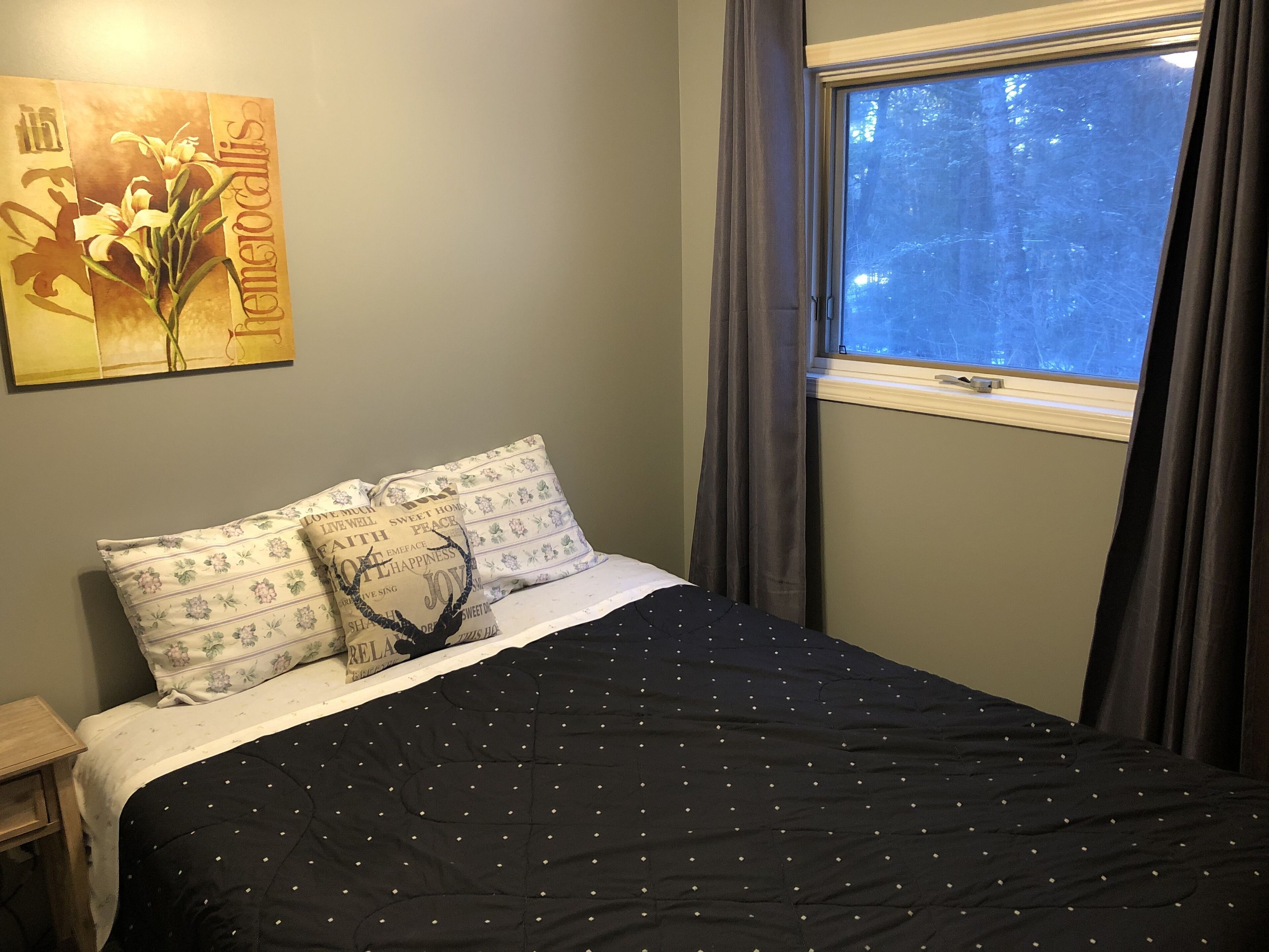Un dormitorio con cama, almohadillas florales, colcha de lunares oscuros, mesita de noche, ventana con cortinas y un cuadro floral en la pared.