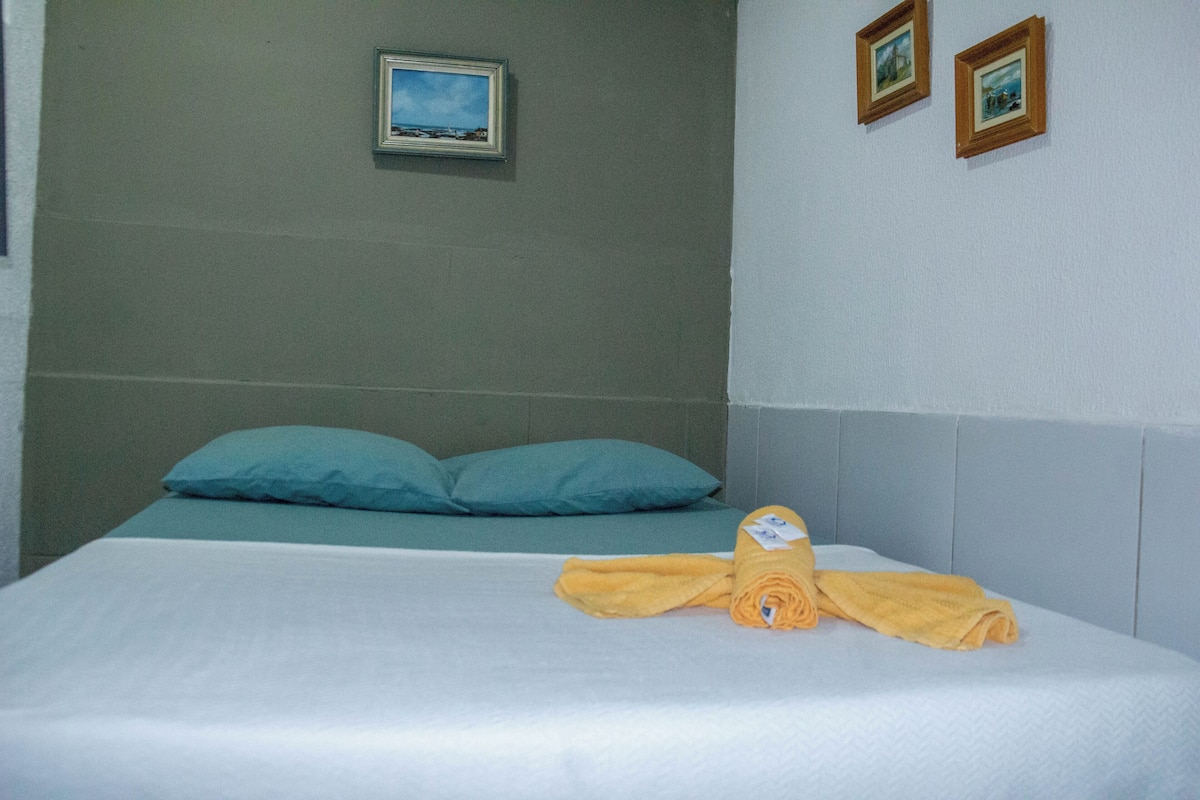 A neatly made bed with a blue pillow, a rolled-up towel, and two framed pictures on the wall.