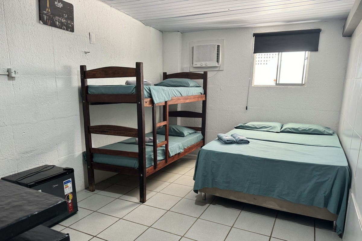 A dormitory room with bunk beds, a desk, a radiator, and a window with a view of buildings.