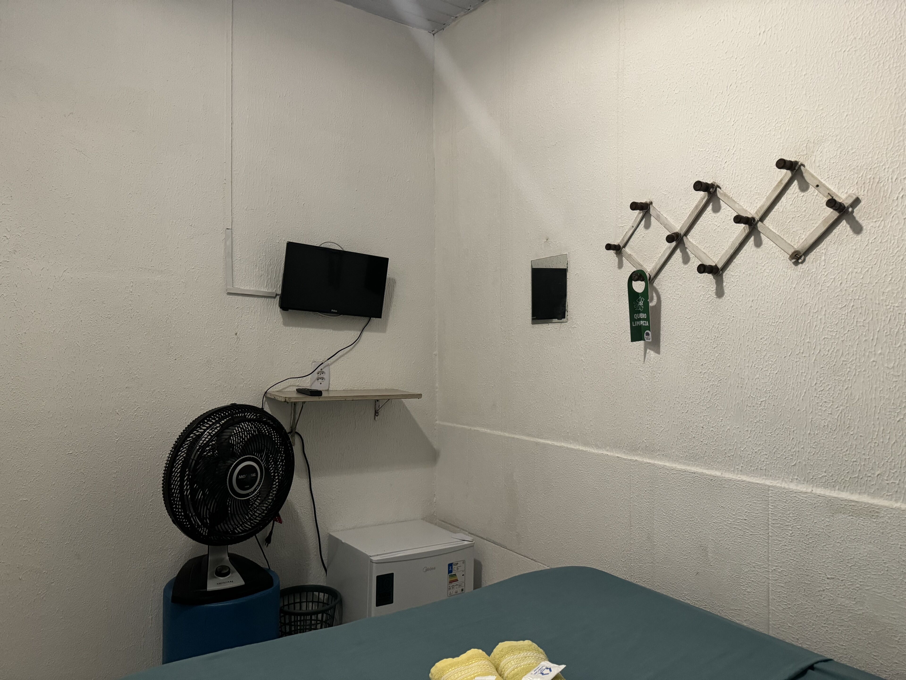 Quarto pequeno com cama, televisão na parede, ventilador e frigobar.