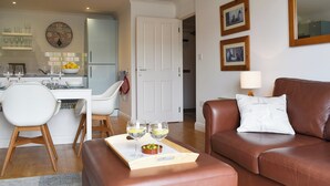 A modern living room with a brown leather sofa, a dining area with a white table and chairs, and a kitchen in the background.