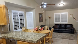 Una cocina con encimera de granito, armarios de madera, lavavajillas, comedor con mesa y sillas de madera, sofá de cuero negro y ventilador de techo.