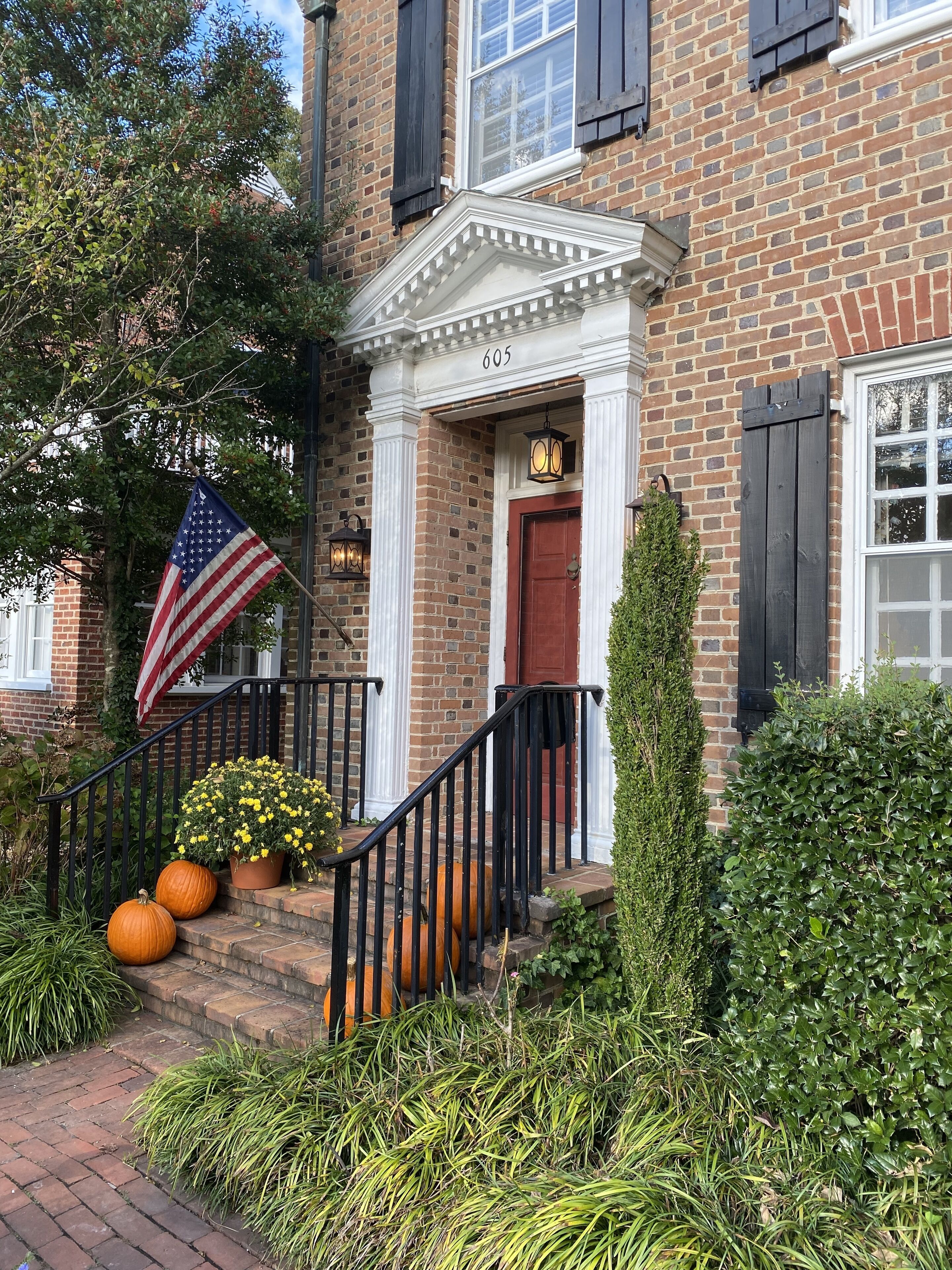 A brick building with a red door, a white porch with a black railing, and an American flag.