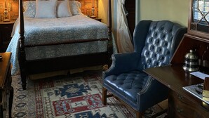 A bedroom with a four-poster bed, a blue upholstered chair, a patterned rug, and a bookshelf with framed pictures. - Inn at 605 (Williamsburg)