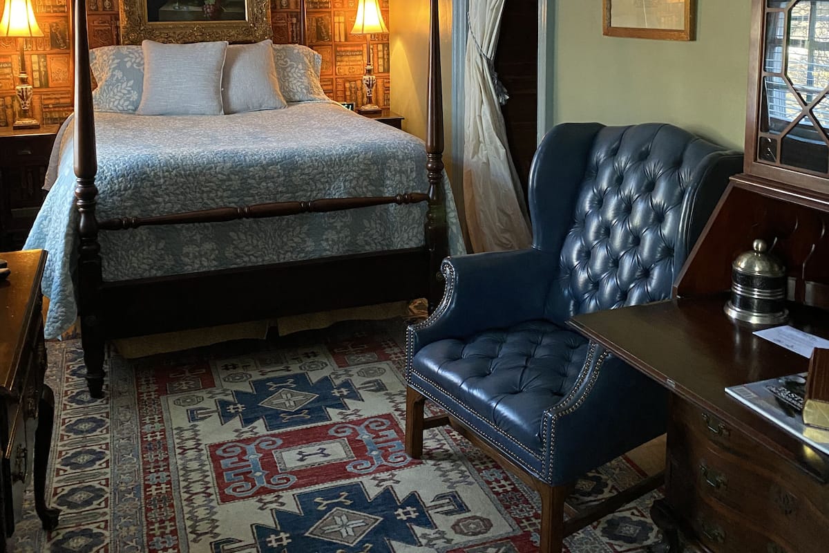 Une chambre à coucher avec un lit à baldaquin, un fauteuil bleu recouvert de tissu, un tapis à motifs et une bibliothèque ornée de cadres avec des photos.