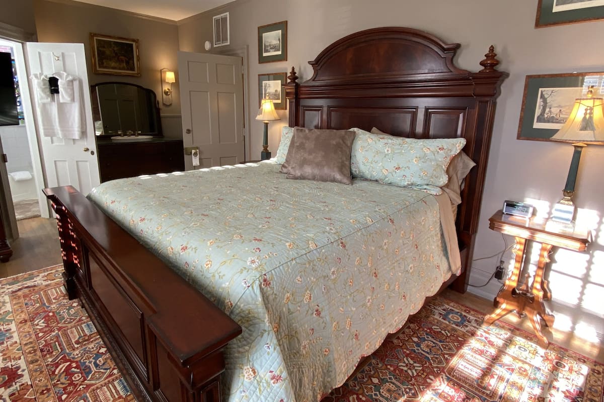 Une chambre avec un grand lit, une tête de lit en bois, une table de chevet avec une lampe, un miroir et une porte sur laquelle sont accrochées des serviettes.