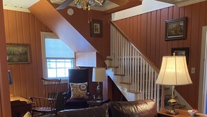 A living room with a brown leather sofa, a ceiling fan, a staircase, and a dining area in the background. - Inn at 605 (Williamsburg)