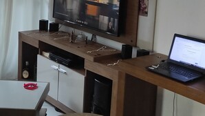 Ein modernes Wohnzimmer mit einem Flachbildfernseher an einer Holzvitrine, einem Laptop auf einem Schreibtisch und einem Essbereich mit Tisch und Stühlen.
