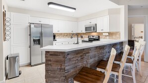 Eine moderne Küche mit weißen Schränken, einem Edelstahlkühlschrank, einer Mikrowelle und einer integrierten Kochinsel mit Barhocker.